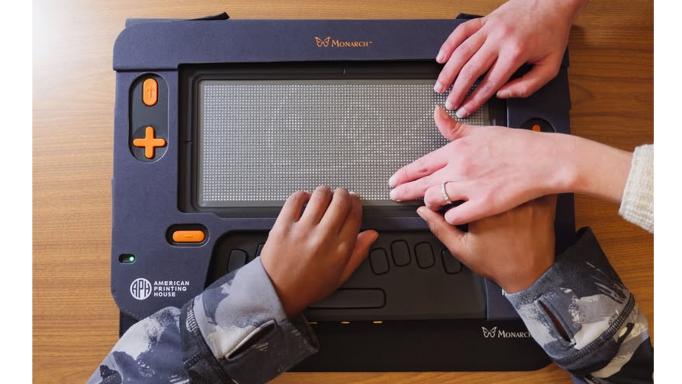 Photo of a child's hands touching the Monarch 2-D Braille display with a woman's hands guiding the child.